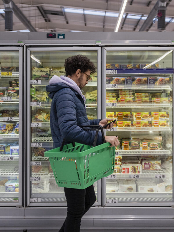 Man shopping in a supermarket while on a budget. He is looking for low prices due to inflation, standing looking at his phone in front of a row of freezers. He is living in the North East of England.